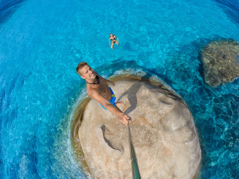 Selfie. Self Portrait On The Top Of The Rock That Surrounds Blue Water. His Girlfriend Is In Water Behind Him. Wide Angle Shot.