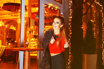 Beautiful young woman poses near bright cafe on the street at night