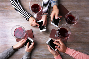 Fototapeta na wymiar Four hands with smart phones holding glasses with red wine, on wooden table background