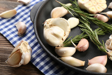 Fresh garlic with rosemary in black pan near blue checkered cotton napkin on wooden background, close up