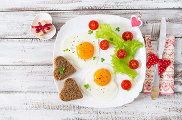 Fototapeta Breakfast on Valentine's Day - fried eggs and bread in the shape of a heart and fresh vegetables. Top view obraz