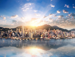 Hong Kong city skyline view from harbor with skyscrapers buildings reflect in water at sunset with sunlight and sun rays shine through clouds on blue sky - obrazy, fototapety, plakaty