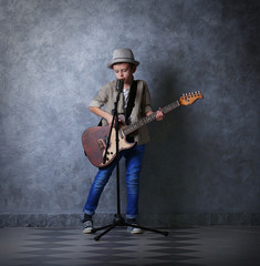 Fototapeta premium Little boy playing guitar and singing with microphone on a grey wall background