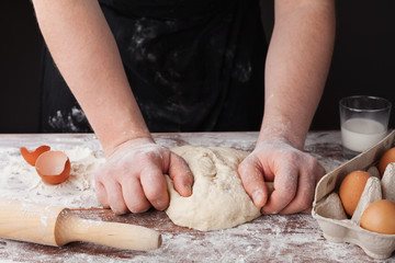 Baker in black apron prepares the dough on a wooden table, male hands knead the dough with flour, homemade dough for bread or pizza, rustic style