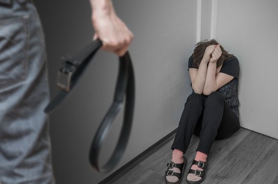 Scared crying and abused woman sitting in corner and fury man with belt. Domestic violence concept.