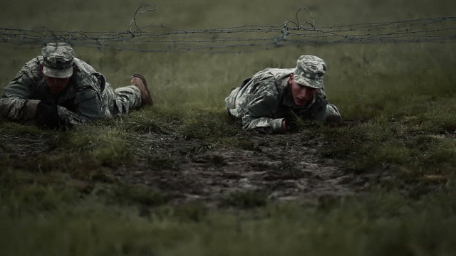 Soldiers crawling under low barbed wire at an obstacle, it's muddy.