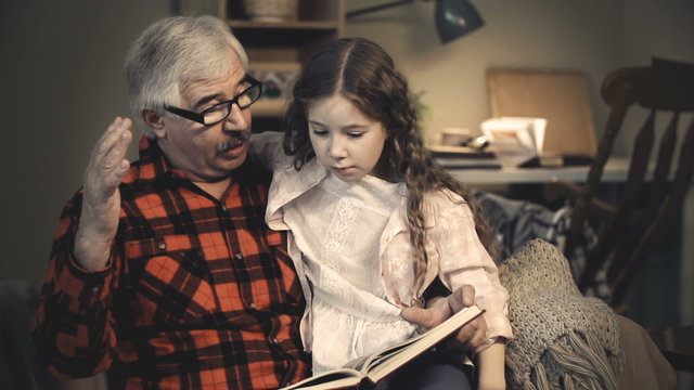 Intelligent grandfather showing his little granddaughter pictures in book and telling interesting facts 