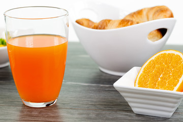 Vitaminreiches Frühstück. Glas Orangensaft und halbierte Orange in eckiger Schale. Weiße Schale mit Hörnchen im Hintergrund. Auf dunklem Holztisch.