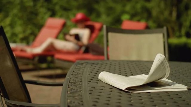 A young woman reading by the pool, with a newspaper on a table in the foreground, in slow motion