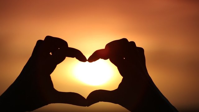 Silhouette of hand in heart shape with sun down in the middle 
