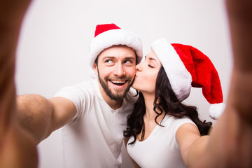 christmas, holidays, technology and people concept - happy couple in santa hats taking selfie picture with smartphone on white background