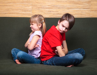 Brother and sister  wearing casual clothes  sitting on a green sofa at home arms crossed back to back angry with each other