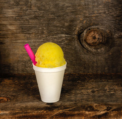 Yellow Hawaiian shaved ice, shave ice or a snow cone with a pink straw in a white cup with a rustic barn wood background.