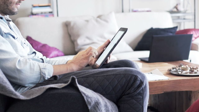 Man hands browsing photos on tablet computer sitting on chair at home
