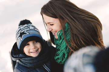 Cute young pregnant mother and her child having fun in winter park on a bright day hugging each other and smiling
