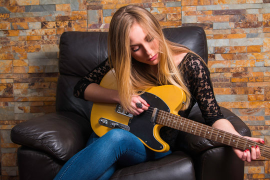      Attractive young girl in blue jeans and black lace shirt sitting in chair and playing guitar on brick background 