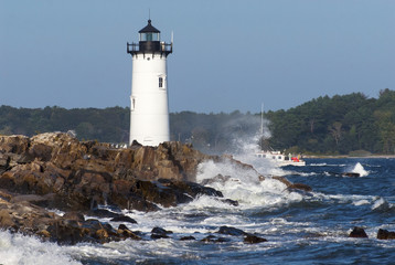 Portsmouth Harbor lighthouse guides fishing boat as waves break over rocky shore during high tide...