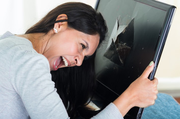 Brunette hispanic model holding up broken black computer screen while smashing her head into it
