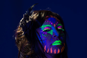 Headshot woman wearing awesome glow in dark facial paint, blue based with other neon colors and obscure abstract background, facing camera
