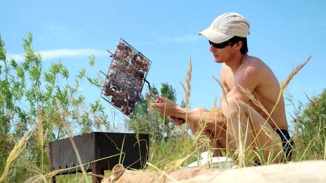 Young man in sunglasses fry the meat on the beach near the sea. Hot food Shashlik in Russia.