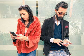 Hipster couple in sad moment ignoring each other using mobile phones - Concept of apathy sadness addicted to new technologies - Boyfriend and girlfriend break up with smartphones addiction
