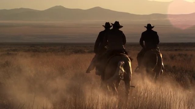 Slow motion shot of Cowboys riding towards distant mountains at dusk with sun flares, golden hour.