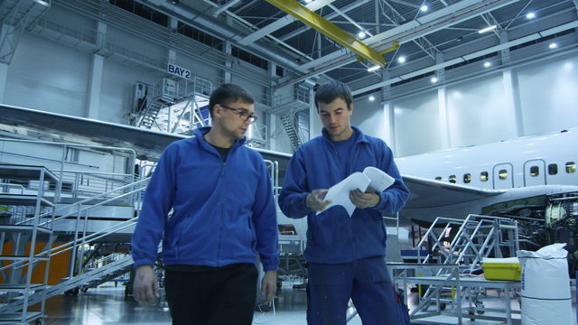 Two aircraft maintenance mechanics have a conversation while checking papers in a plane hangar with an airplane in the background. Shot on RED Cinema Camera.