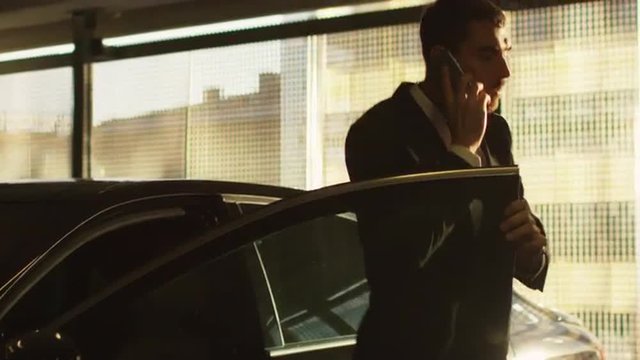 Businessman gets out of a black executive car at sunset while speaking on a phone. Shot on RED Cinema Camera.