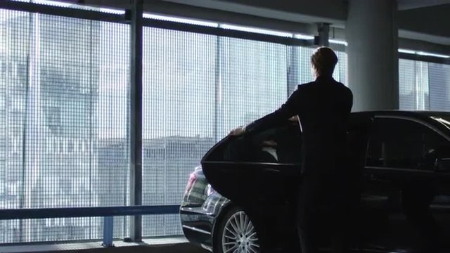 Driver opens the door for a businessman who speaks on the phone to step out of the black executive car at a garage parking.