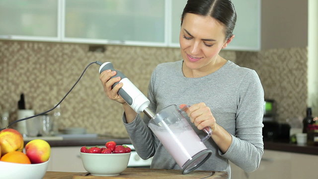 Cheerful brunette woman cooking dairy beverages with electric blender from strawberries at domestic kitchen