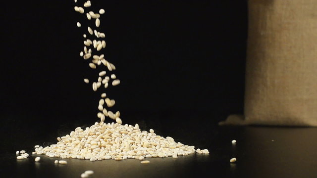 SLOW (240fps): A pearl barlies fall in a pile on a table. A cloth bag stands on a background
