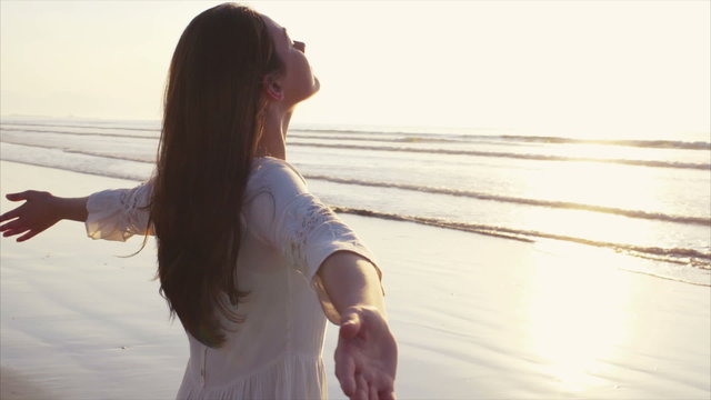 Tilt up video of young woman standing on shore. Side view of carefree female with arms outstretched and eyes closed. Woman in white sundress enjoying nature on beach during sunset.