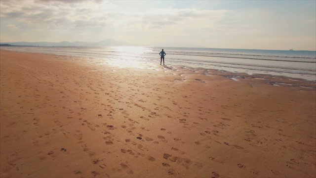 Aerial drone footage of fit woman standing on shore. Tracking shot of idyllic beach on sunny day. Rear view of female standing with hands on hips. Video is filmed from high angle perspective.