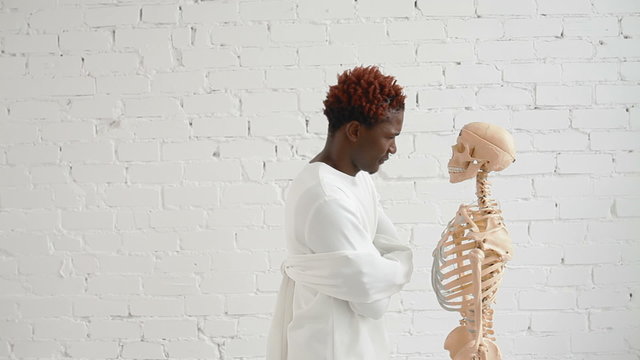 An insane black man wearing a straitjacket dance and looking at human skeleton stand