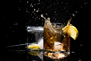 Falling piece of ice in quality glass of whiskey and makes very many splashes and another glass lying on side with lemon slice inside, tubules and ice on black background.
