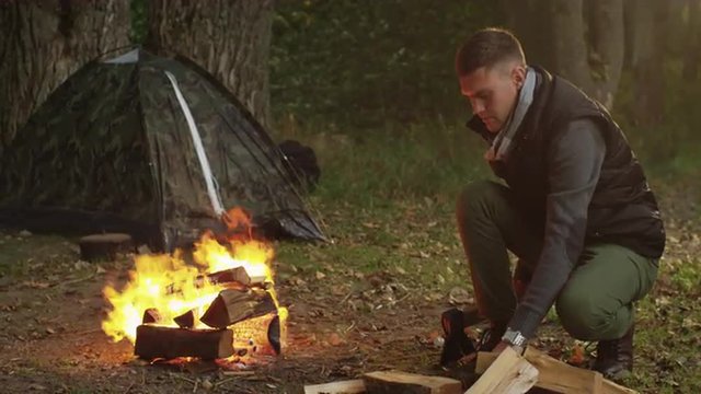 Short haired man in autumn clothes is chopping wood with an axe and throwing it into the fire. Shot on RED Cinema Camera.
