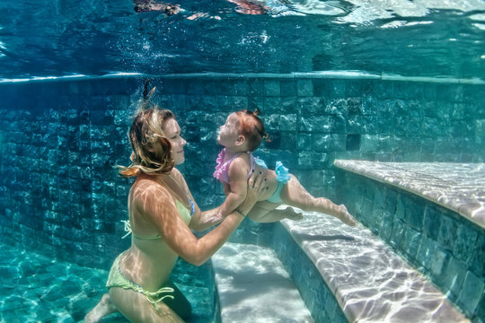 Happy family - mother hold in hands baby girl and dive down with fun in beach pool, active parents lifestyle, people water sport activity and underwater swimming lesson on summer vacation with child.