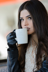 Woman with a unique fleshy lips wearing leather jacket and gloves holding a coffee cup, sitting in cafeteria