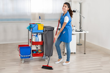 Happy Female Janitor Cleaning Floor With Broom In Office
