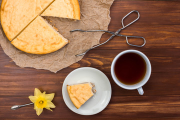 New York cheesecake on rustic wooden background with cup of tea, tongs for dessert and yellow daffodil. Biting a piece of cheesecake. 