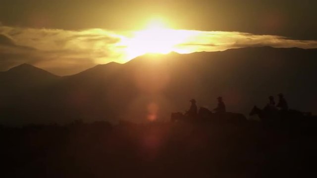 Silhouetted cowboys riding at sunset, kicking up dust. Shot in slow motion with a lens flare.