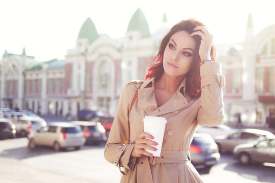 Early coffee / Beautiful young woman in a modern trench coat, holding a disposable takeaway cup and standing against urban city background.