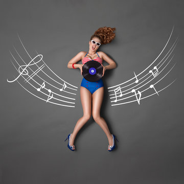Sitting on a staff / Beautiful pin-up girl in retro bikini and sunglasses, holding an LP microgroove vinyl record and sitting on musical staff chalk drawings background, top view.