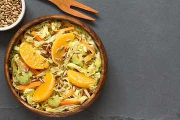 Fresh salad made of savoy cabbage, carrot, celery, and orange with roasted sunflower seeds on top, photographed overhead on slate with natural light (Selective Focus)