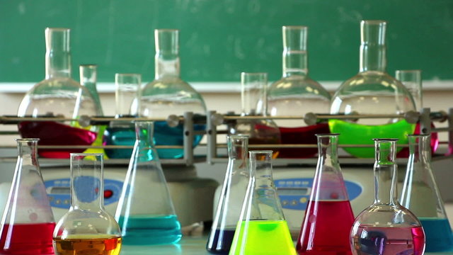 Laboratory glasswares filled with colorful solutions on the rotating shakers and green chalkboard background. Focus on the front row of flasks standing. Lockdown.