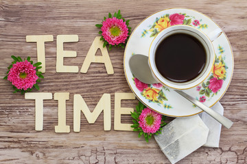 Tea time written with wooden letters, tea in vintage cup, tea bags and pink daisies
