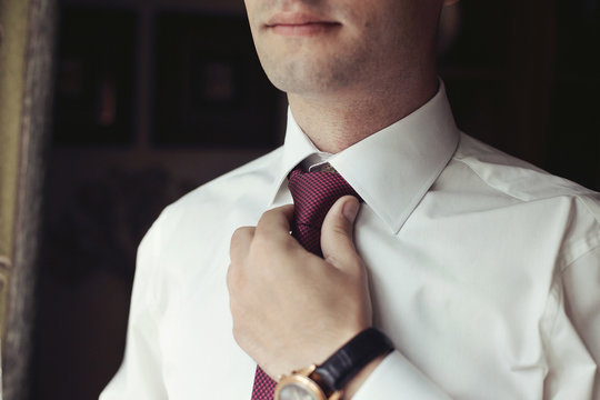 Handsome Groom Preparing For Wedding, Putting On Tie Near Window