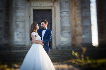 Sensual romantic newlywed bride and groom hugging in front of old baroque church with columns at sunset