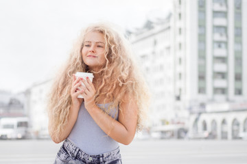 Happy smile / Happy smile. Happy young woman with long curly hair, holding a take away coffee cup and smiling against city background.