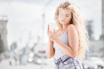 Happy smile / Happy young woman with long curly hair, holding a take away coffee cup and smiling with flirt in front of a camera against urban city traffic background.